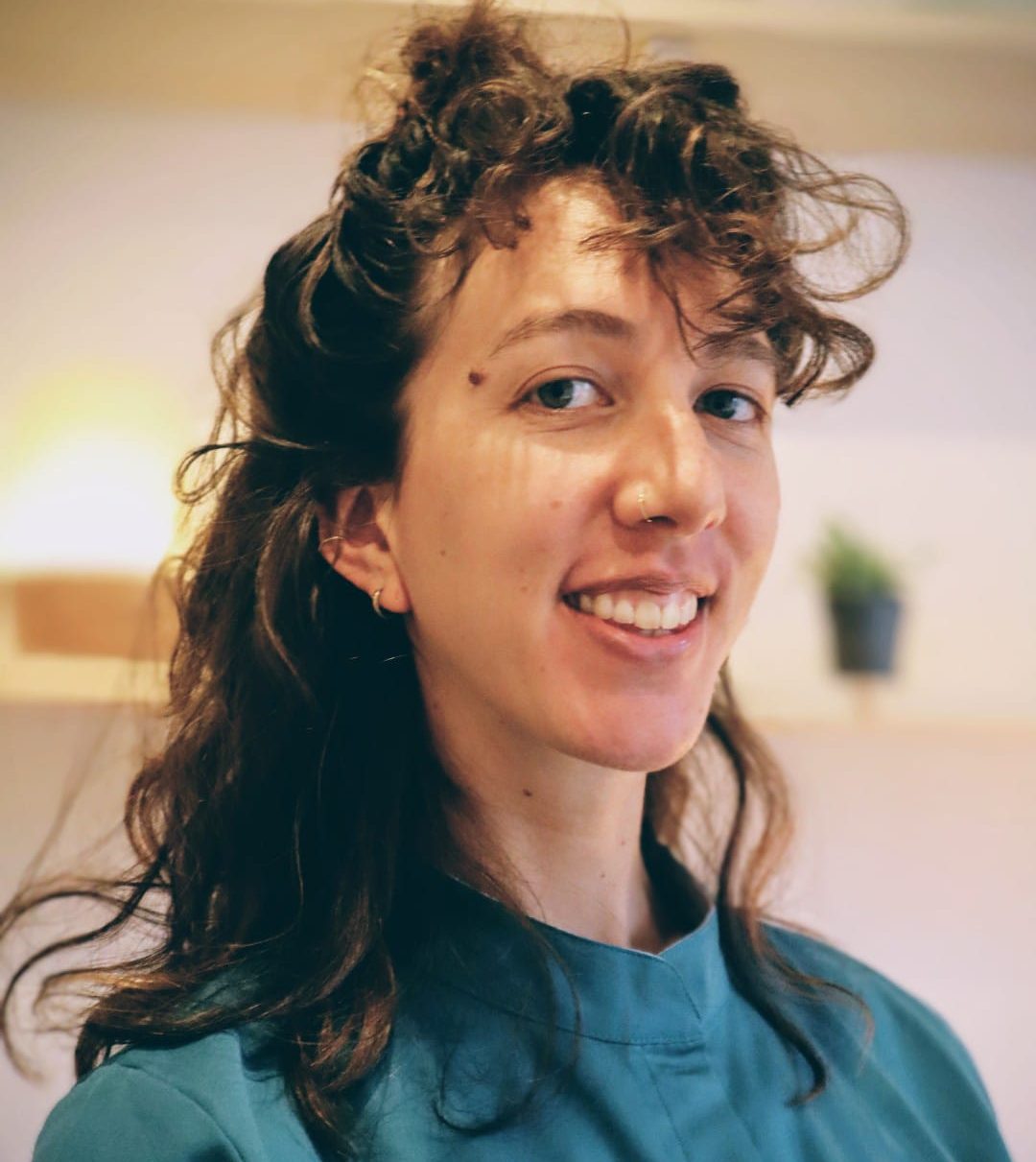 Photo de présentation Femme souriante avec des cheveux frisés, portant un haut bleu, dans un intérieur lumineux. Photo présentant la praticienne de massage