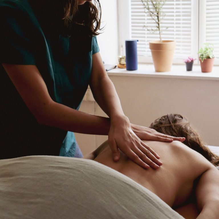 Massage du dos Femme recevant un massage, avec une praticienne en arrière-plan. Plantes en pot visibles.